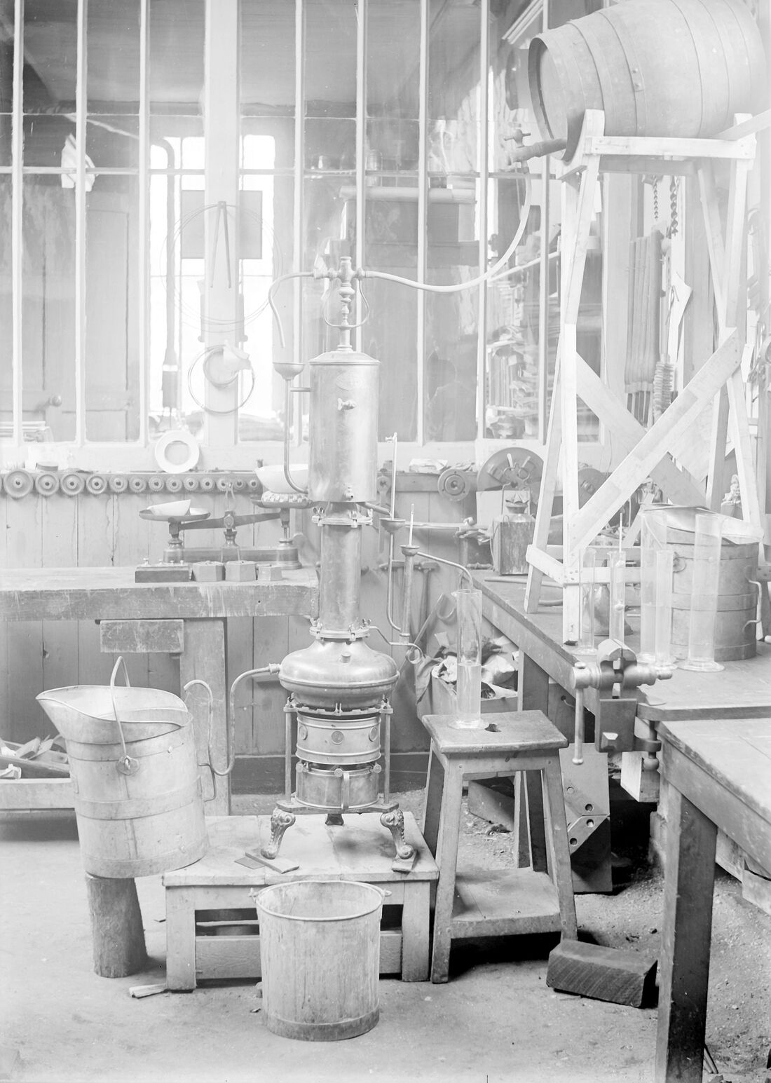 Photographie noir et blanc d'un atelier : un petit alambic de cuivre à colonne, monté sur un réchaud, entouré d'une balance à plateaux, d'éprouvettes graduées, d'un seau de bois et d'un fût sur chevalet.