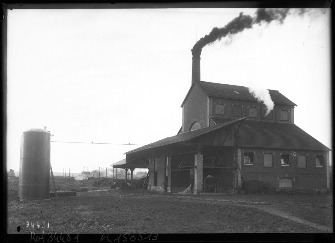 Photographie noir et blanc d'une distillerie industrielle en activité : bâtiment de brique à deux étages, halle couverte adossée, haute cheminée crachant une fumée noire, cuve cylindrique isolée à gauche.