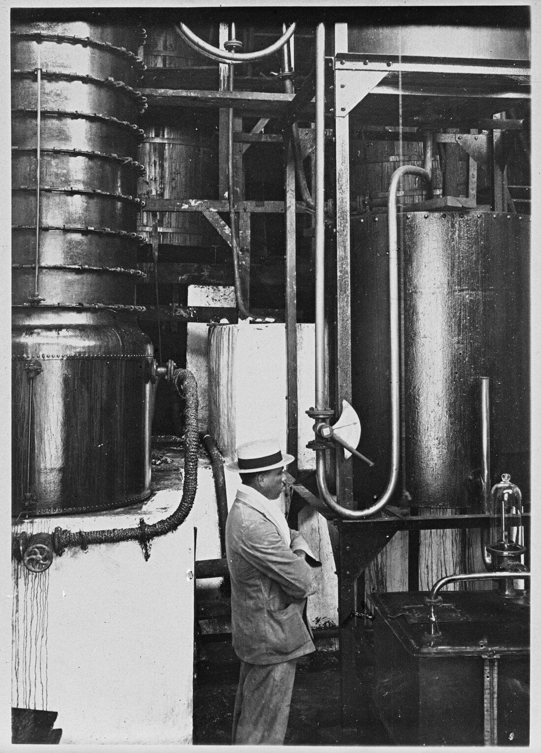 Photographie noir et blanc : l'intérieur d'une distillerie de rhum, colonnes et cuves métalliques ; un homme en costume clair et chapeau observe les appareils.